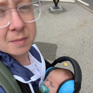 A selfie of Alex, a white man with red hair wearing clear framed glasses carrying Alice, a white baby with brown hair wearing light blue and black ear defenders. 