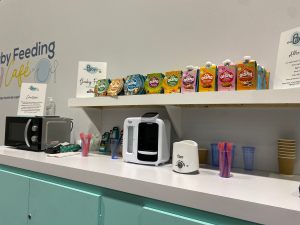 A photo of a baby foodprep area with microwave, prep machine, bottle warmer and cups with pink and purple spoons in. Above is a shelf with various coloured bowls and pouches containing meals for babies. 